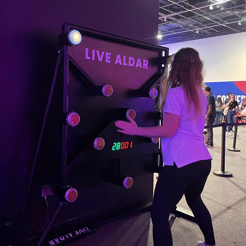 Woman playing Batak reaction game at a fitness event in Dubai, touching illuminated buttons to test her speed and reflexes. "Speedy Speedy" displayed on wall.