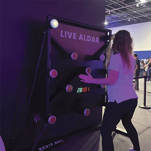 Woman playing Batak reaction game at a fitness event in Dubai, touching illuminated buttons to test her speed and reflexes. "Speedy Speedy" displayed on wall.