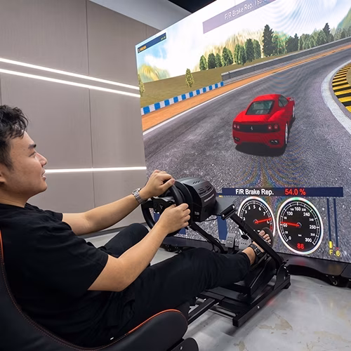 Woman using a pro-grade racing simulator setup with a high-precision wheel, pedals, black bucket seat, and large video screen showing a sports car track experience.