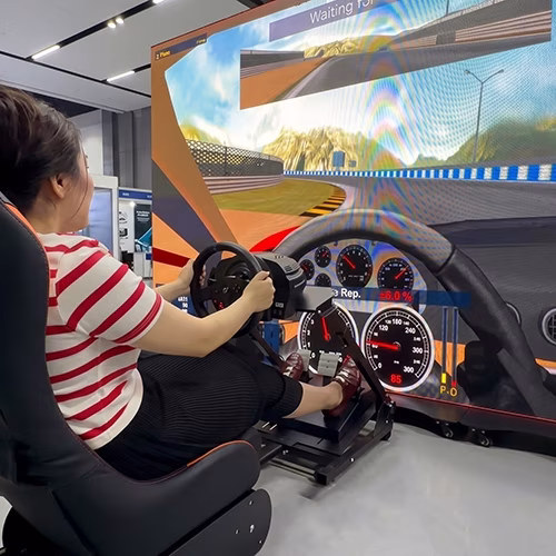 Woman using a pro-grade racing simulator setup with a high-precision wheel, pedals, black bucket seat, and large video screen showing a sports car track experience.