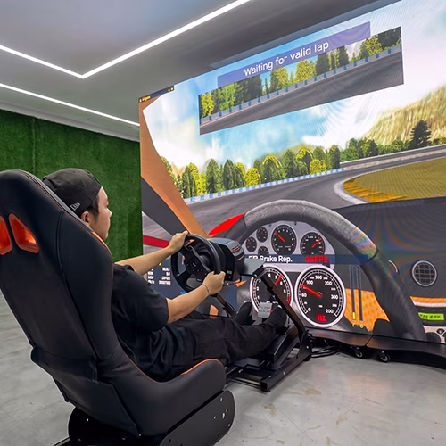 Woman using a pro-grade racing simulator setup with a high-precision wheel, pedals, black bucket seat, and large video screen showing a sports car track experience.