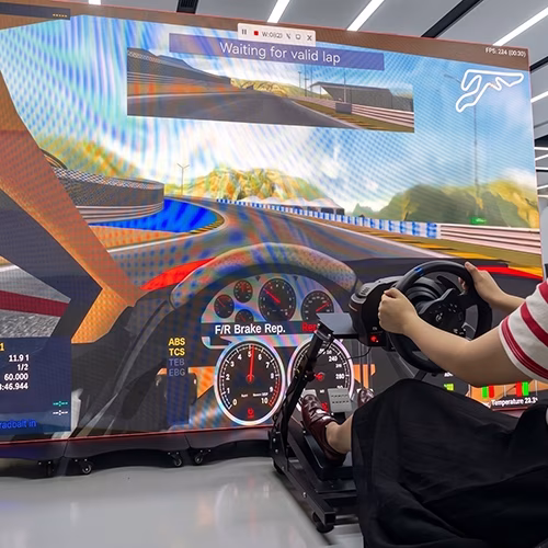 Woman using a pro-grade racing simulator setup with a high-precision wheel, pedals, black bucket seat, and large video screen showing a sports car track experience.