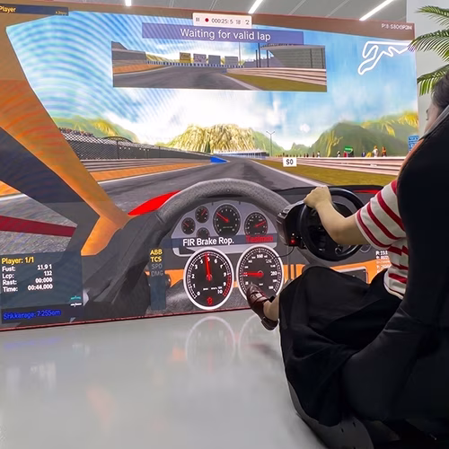 Woman using a pro-grade racing simulator setup with a high-precision wheel, pedals, black bucket seat, and large video screen showing a sports car track experience.