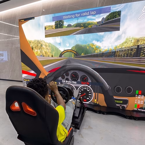 Woman using a pro-grade racing simulator setup with a high-precision wheel, pedals, black bucket seat, and large video screen showing a sports car track experience.