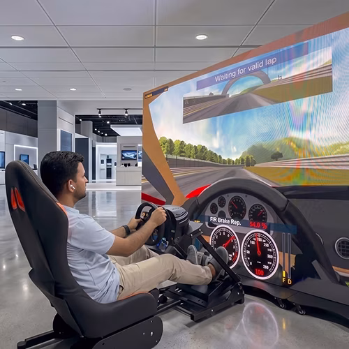 Woman using a pro-grade racing simulator setup with a high-precision wheel, pedals, black bucket seat, and large video screen showing a sports car track experience.