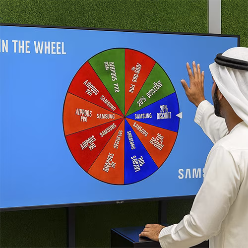 Arab male playing Spin the Wheel game at an exhibition event in Dubai, wearing traditional kandura, fully branded setup with digital screen and prize display, engaging crowd.