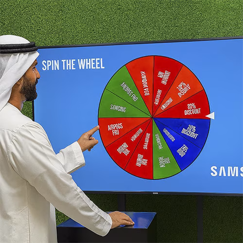 Arab male playing Spin the Wheel game at an exhibition event in Dubai, wearing traditional kandura, fully branded setup with digital screen and prize display, engaging crowd.