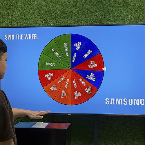 Arab male playing Spin the Wheel game at an exhibition event in Dubai, wearing traditional kandura, fully branded setup with digital screen and prize display, engaging crowd.