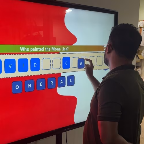 Guest playing word scramble game on large touchscreen at Dubai event with branded graphics and green artificial grass wall background.