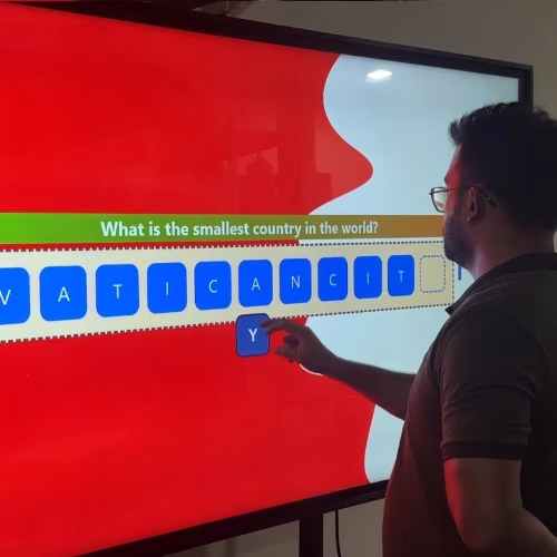 Guest playing word scramble game on large touchscreen at Dubai event with branded graphics and green artificial grass wall background.