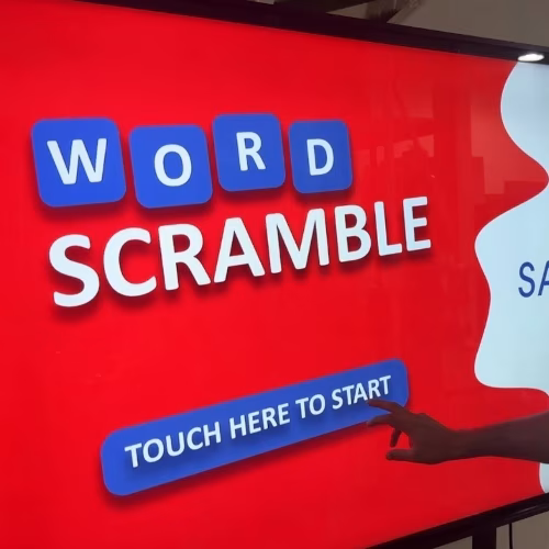 Guest playing word scramble game on large touchscreen at Dubai event with branded graphics and green artificial grass wall background.